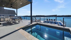 Area piscina all'aperto con solarium in legno, angolo barbecue e poltrone relax con vista sull'acqua.