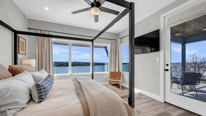 Camera da letto con un letto grande, una ventola a soffitto e vista su un lago attraverso ampie finestre.