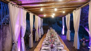 Ein Essbereich im Freien mit einem langen Holztisch, gedeckt für eine Mahlzeit, umgeben von weißen Vorhängen und beleuchtet durch Lichterketten.