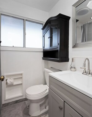 Un baño con inodoro blanco, tocador con espejo y ventana sobre el inodoro.