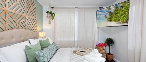 A modern bedroom with a tufted headboard, a large bed with multiple pillows, a flat-screen TV on the wall, a potted plant, and a wooden tray on the bed.