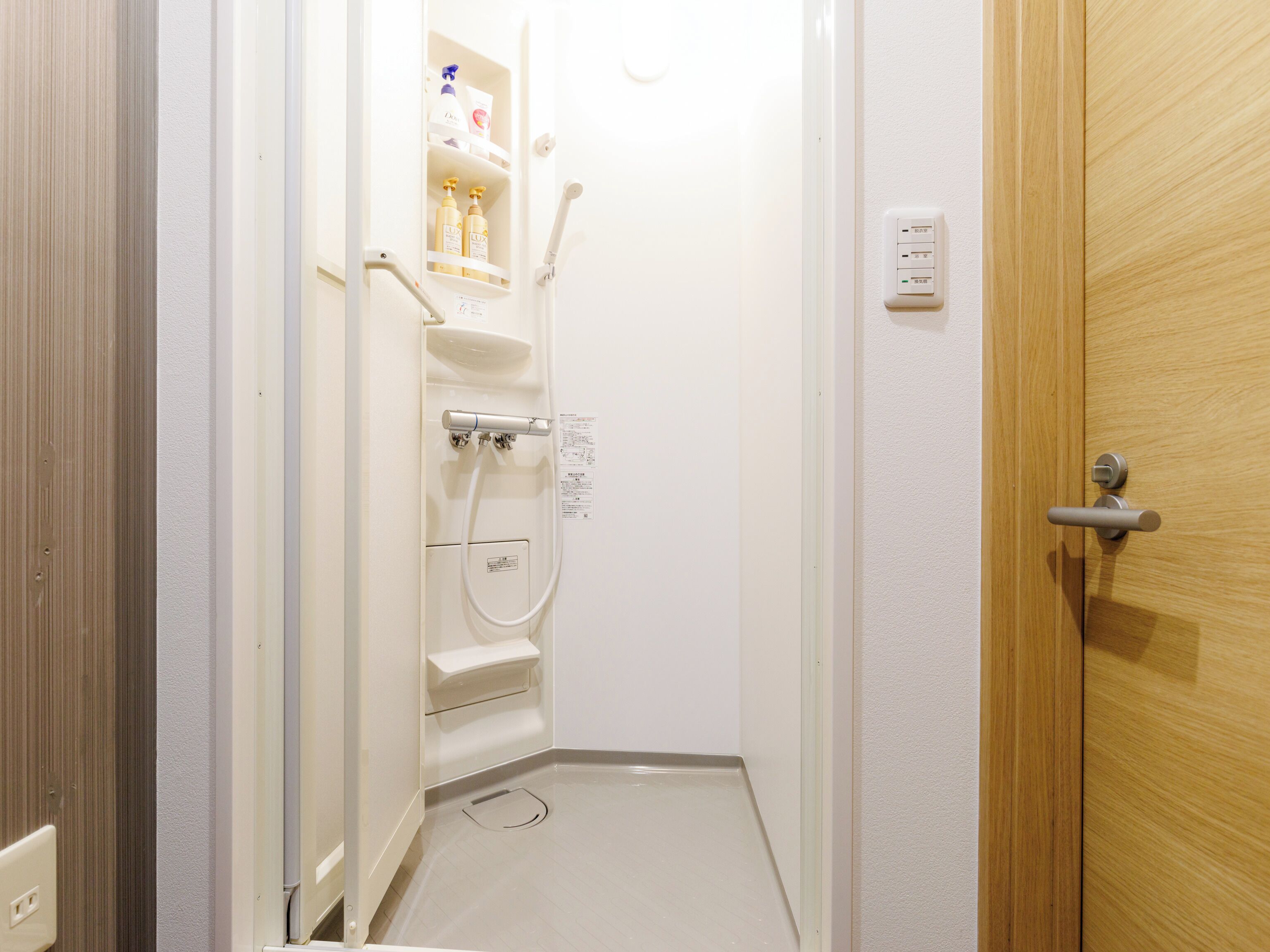 A small, narrow bathroom with a shower, toilet, and sink. There are toiletries on the shelves, and a wooden door is visible.