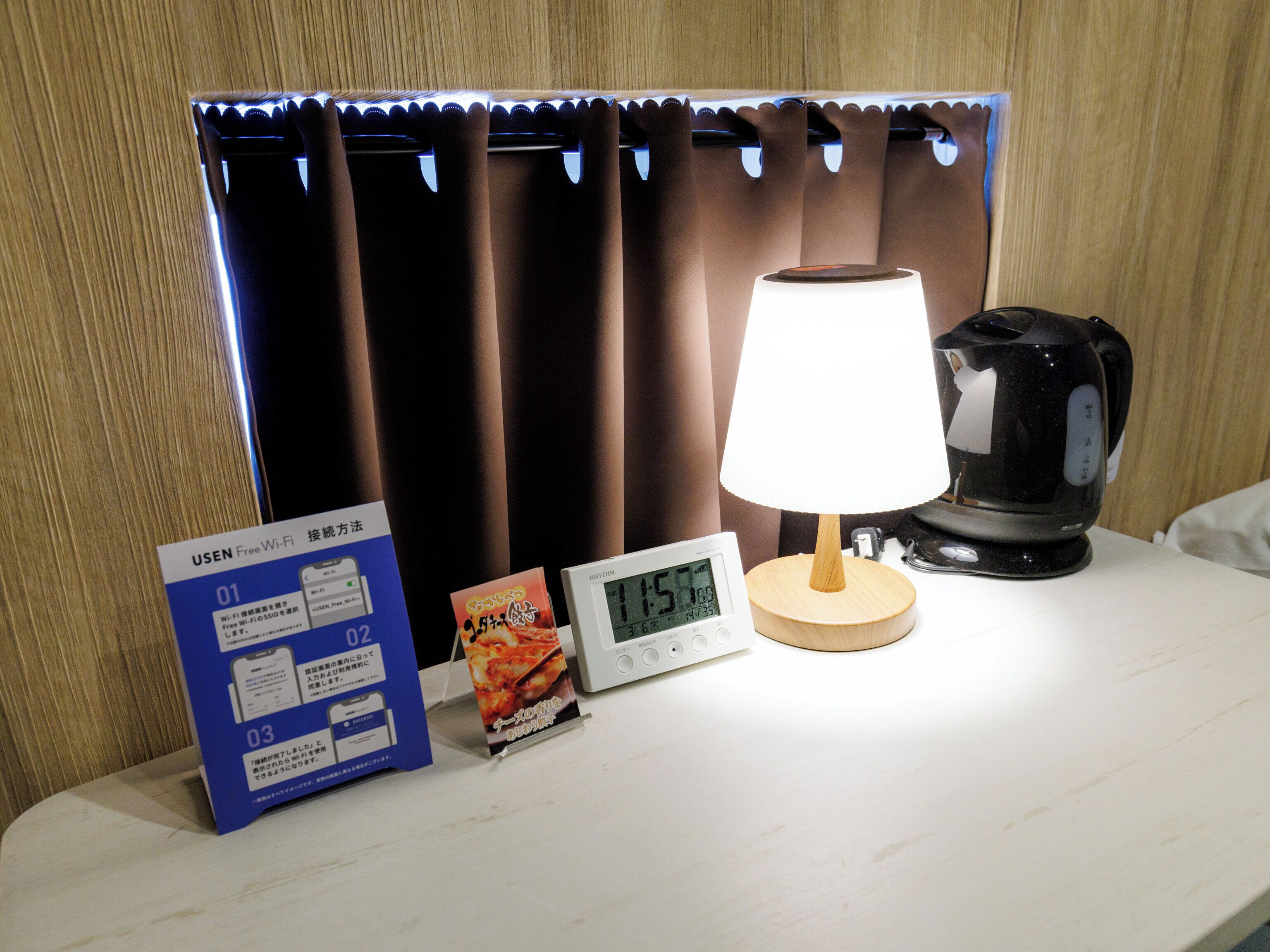 Habitación de hotel con mesita de noche que incluye un reloj digital, una lámpara, un hervidor de agua y folletos informativos.