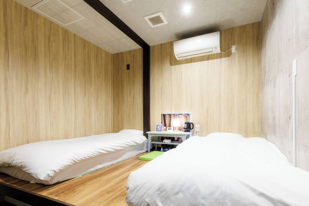 Ein kleines Hotelzimmer mit einem Bett für eine Person, zwei Einzelbetten, einem Holzboden, einem kleinen Schreibtisch mit Lampe, einer Kaffeemaschine und einer Klimaanlage an der Wand.