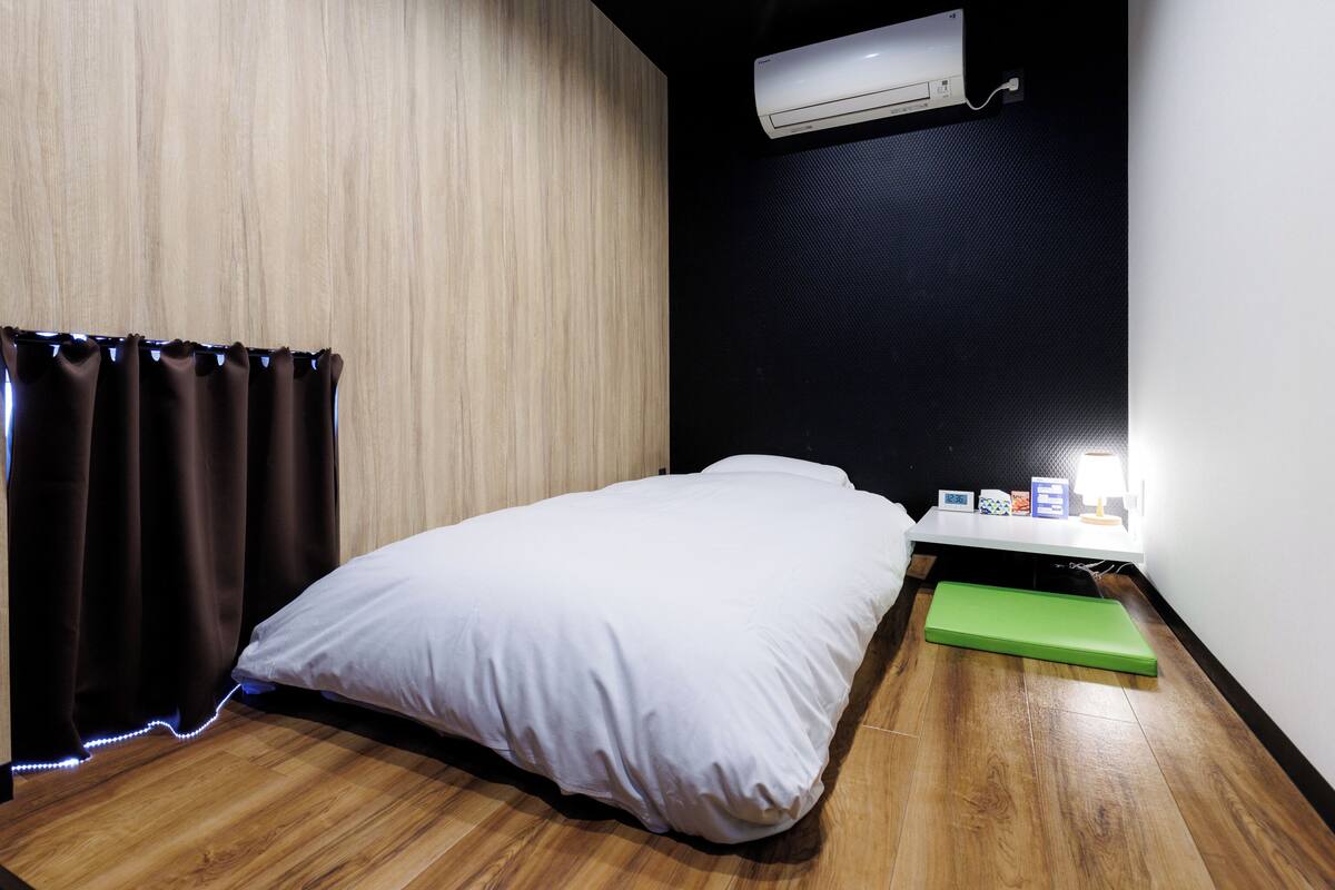 Ein Zimmer mit einem Bett, einem kleinen Tisch mit einer Lampe, einer grünen Matte und einer Klimaanlage.