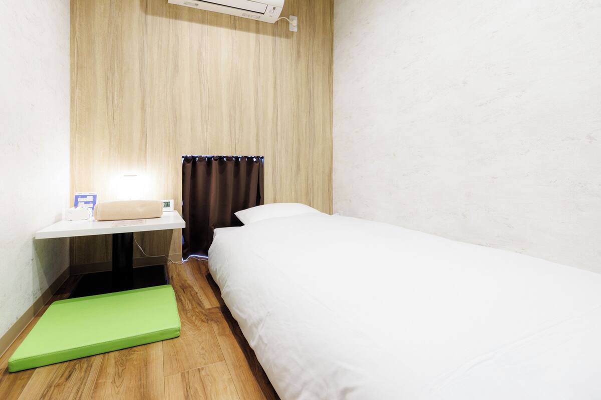 Ein kleines Hotelzimmer mit einem Bett, einem Nachttisch mit Lampe, einer grünen Matte, einem Fenster mit Vorhängen und einer Klimaanlage.