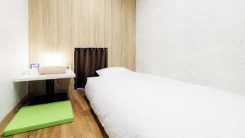 A small hotel room with a bed, a bedside table with a lamp, a green mat, a window with curtains, and an air conditioning unit.