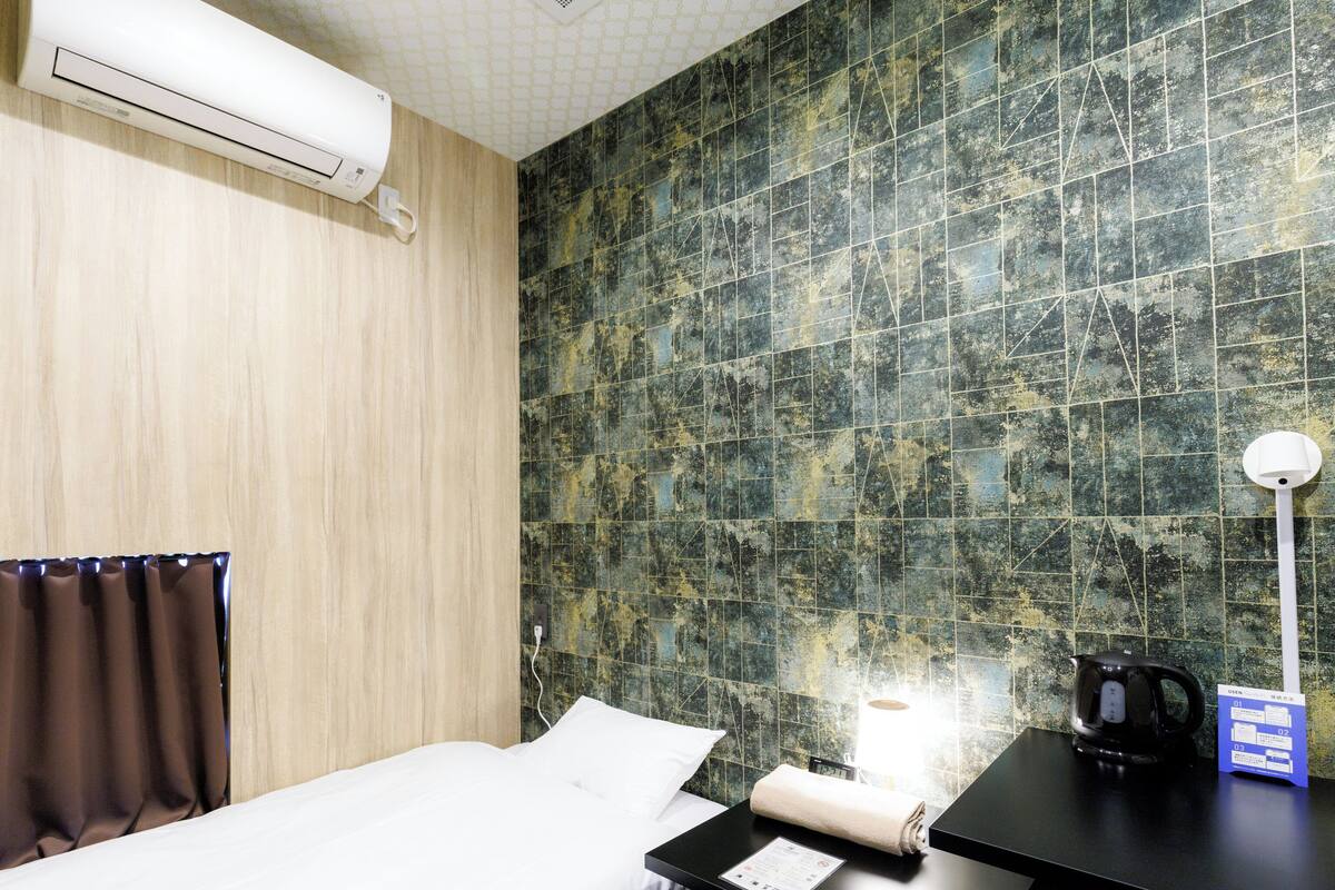 Ein Hotelzimmer mit einem Bett, einem Schreibtisch mit Wasserkocher, einer Lampe und einer Wand mit strukturierter Oberfläche.