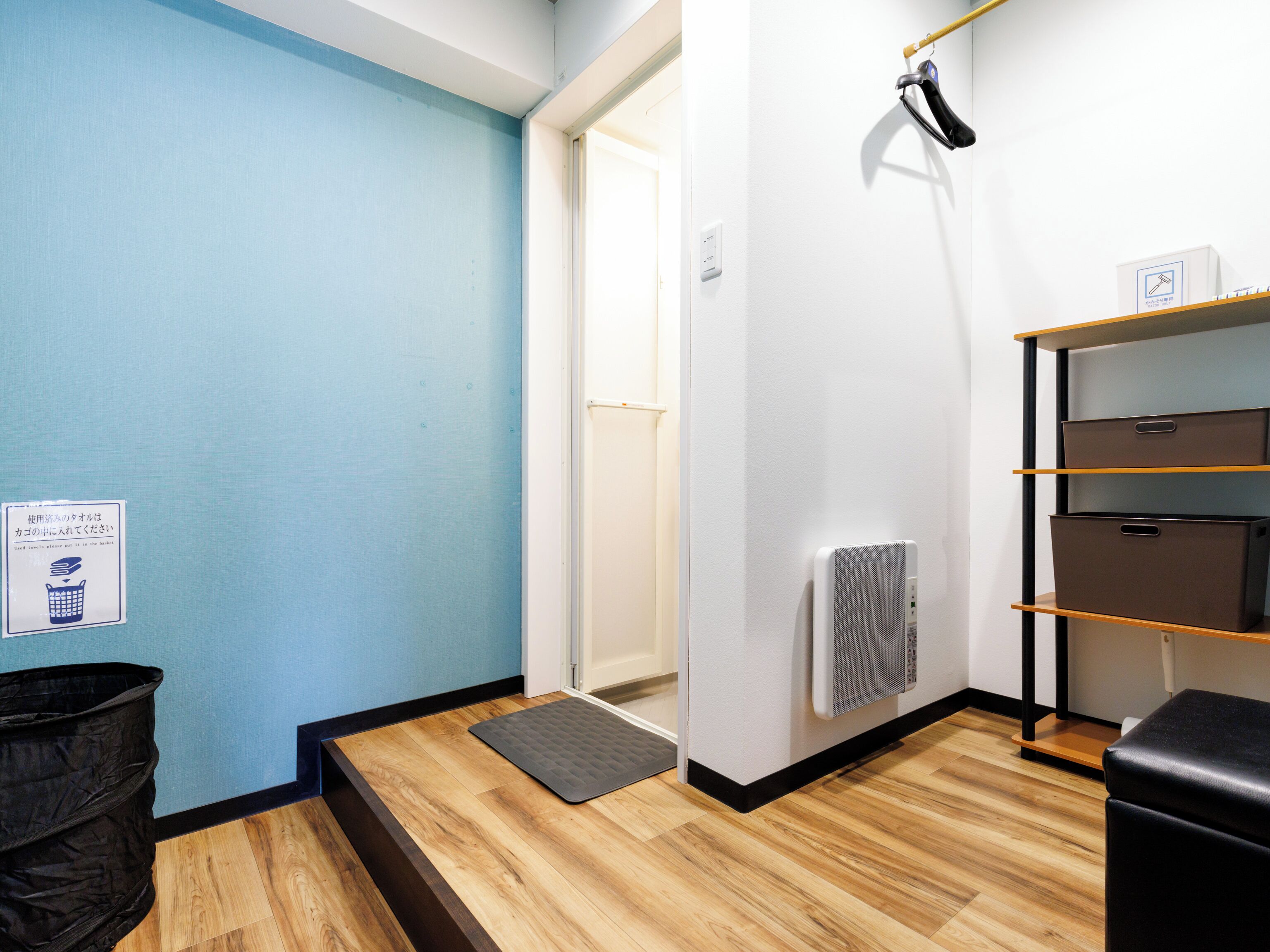 Habitación de hotel con pared azul, suelo de madera, un cubo de basura negro, un banco negro y un estante con cajas de almacenamiento.