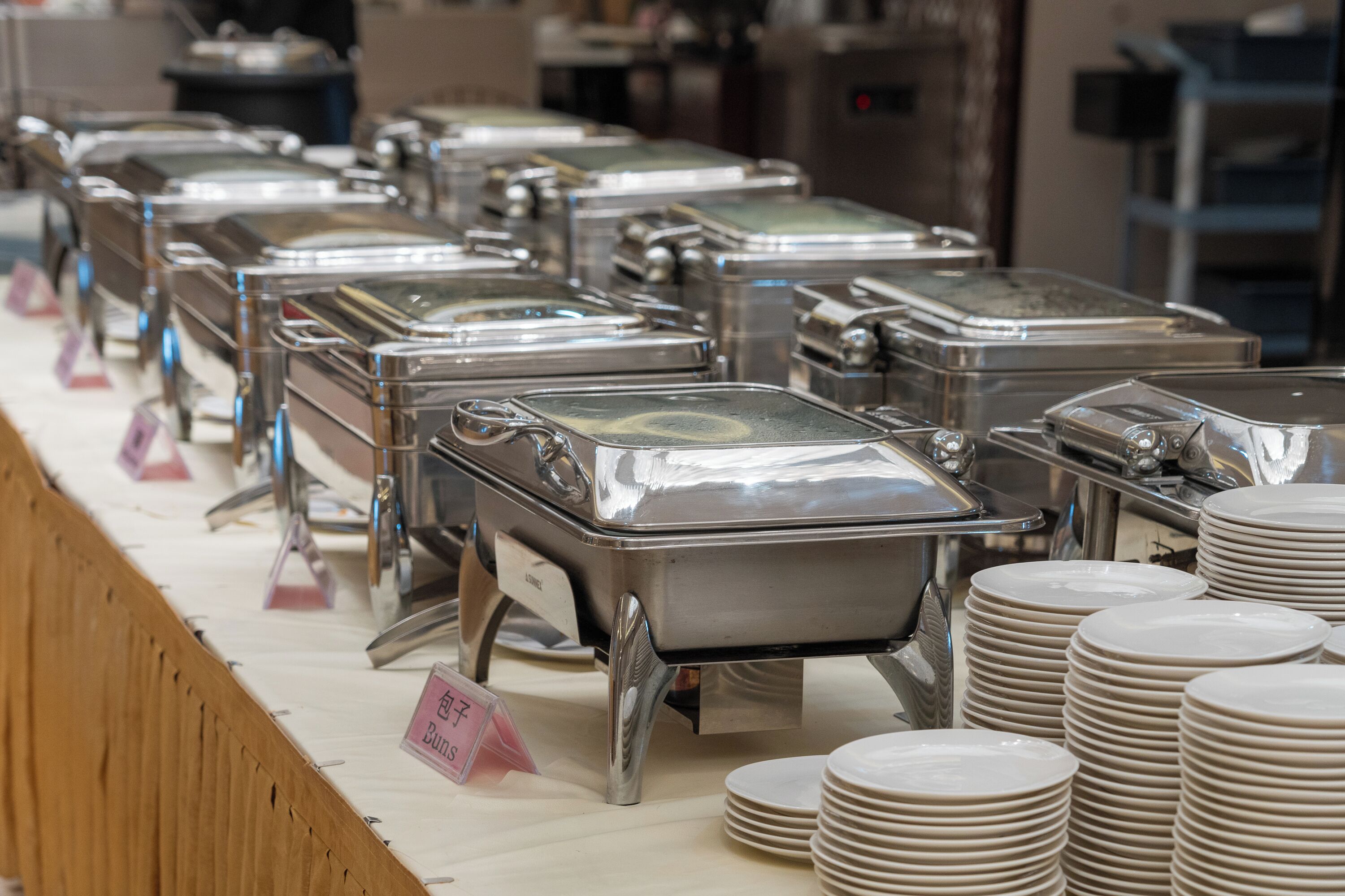 A buffet setup with multiple stainless steel chafing dishes and stacks of white plates.