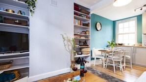 A living room with a TV, shelves with various items, a dining area with a table and chairs, and a kitchen area with a window.
