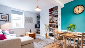 A cozy living room with a sofa, a coffee table, and a dining area with a table and chairs. There is a bookshelf filled with books and decorative items, and a wall clock.
