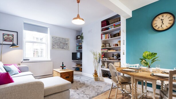 A cozy living room with a sofa, a dining table with chairs, a bookshelf, and a wall clock.