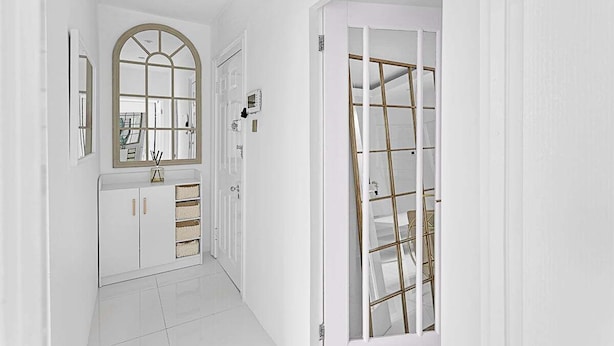 Un couloir aux murs blancs, avec un sol carrelé de blanc, une porte blanche dotée d’un cadre doré et un meuble blanc muni de tiroirs et de paniers.