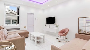 Un salon moderne avec un canapé blanc, une table basse blanche, un fauteuil à bascule rose et un téléviseur à écran plat fixé à un mur blanc.