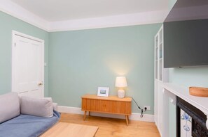 A room with a light green wall, a white door, a blue sofa, a wooden side table with a lamp, and a fireplace with a TV above it. - Traditional Bath House, Garden with City Views (Bath and North East Somerset)