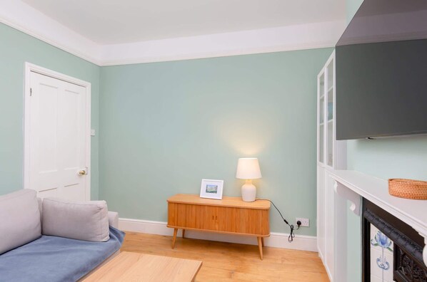 A room with a light green wall, a white door, a blue sofa, a wooden side table with a lamp, and a fireplace with a TV above it. - Traditional Bath House, Garden with City Views (Bath and North East Somerset)