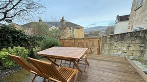 A wooden deck with a table and chairs, surrounded by a stone wall and vegetation. - Traditional Bath House, Garden with City Views (Bath and North East Somerset)