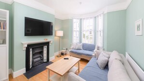 A living room with a fireplace, a TV mounted on the wall, a sofa, a table, and a window with blinds. - Traditional Bath House, Garden with City Views (Bath and North East Somerset)