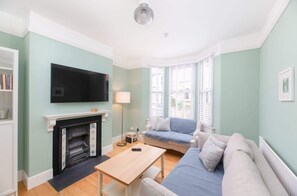 Un salon avec une cheminée, un téléviseur fixé au mur, un canapé, une table et une fenêtre avec des stores.