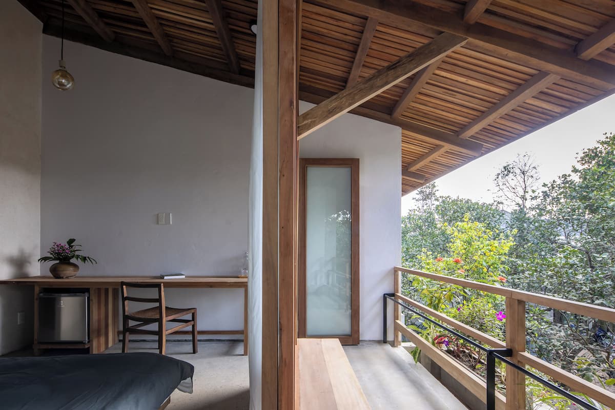 A bedroom with a bed, a desk, and a chair. There is a balcony with a wooden railing and a view of trees and plants.