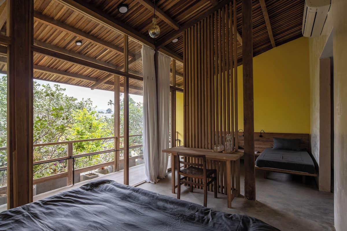 A room with a bed, a table, and a chair. There is a window with curtains, and a view of trees outside.