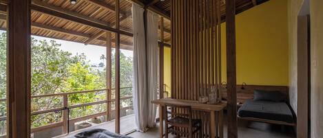 A room with a bed, a table, and a chair. There is a window with curtains, and a view of trees outside.