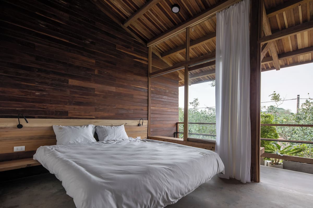 A bedroom with a wooden ceiling, a large bed with white bedding, and a view of greenery through a window.
