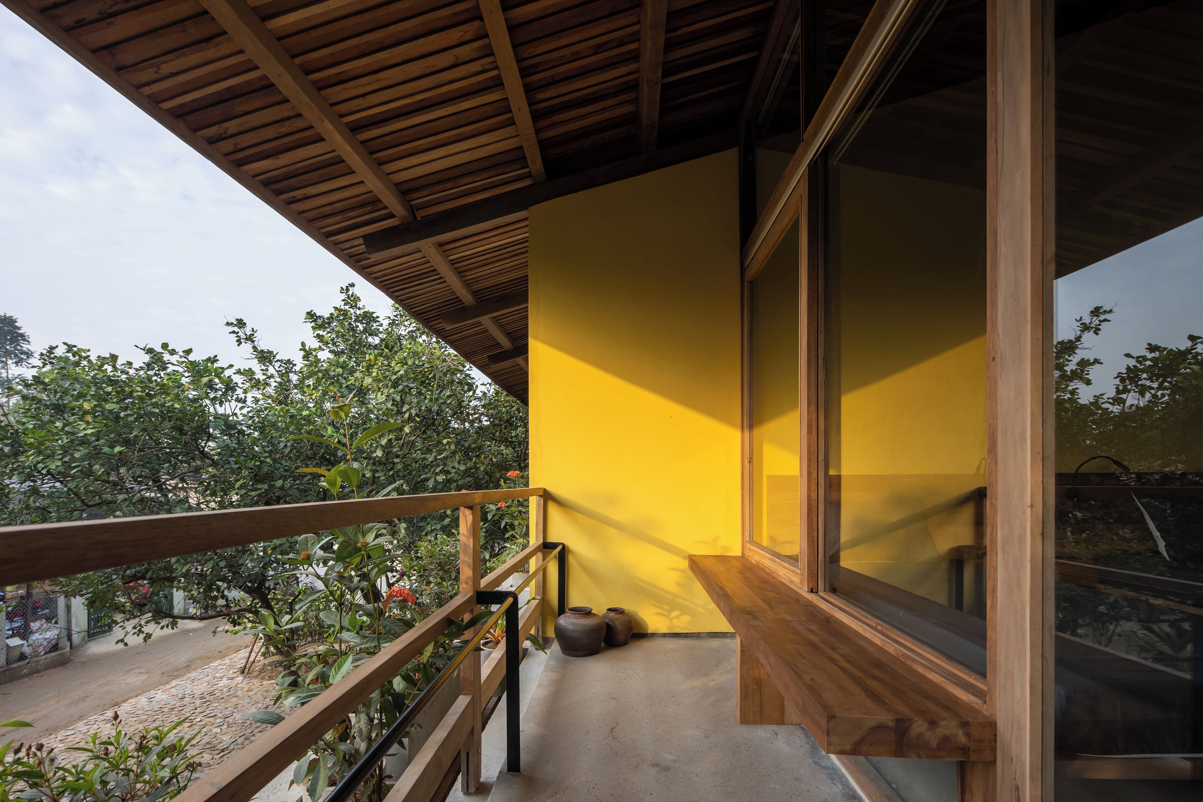 A balcony with a wooden railing, a yellow wall, and a view of trees and a fence.
