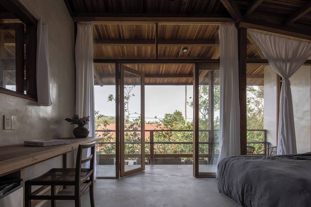 A bedroom with a bed, a desk, and a chair. There is a balcony with a view of trees and buildings.