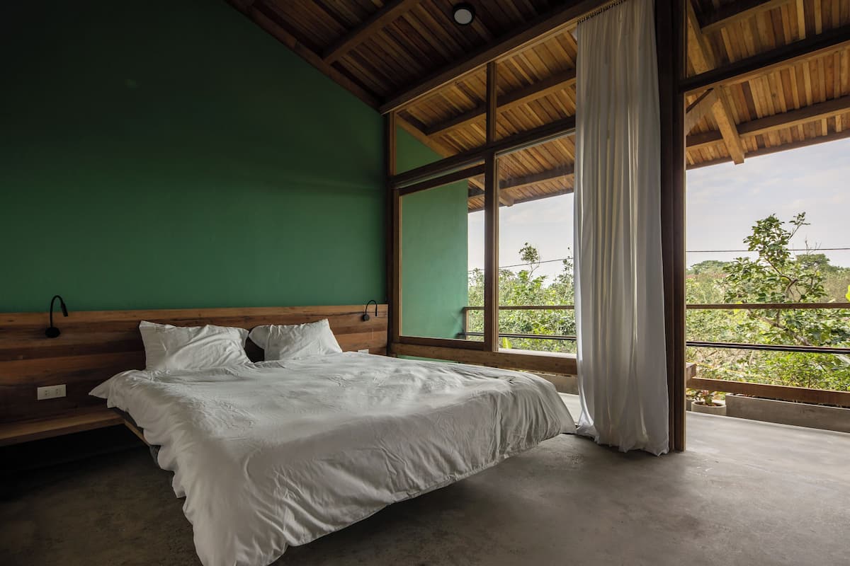 A bedroom with a large bed, wooden headboard, green walls, and a view of greenery through large windows.