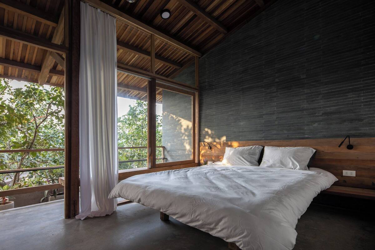 A bedroom with a large bed, wooden ceiling, and a view of trees through large windows.