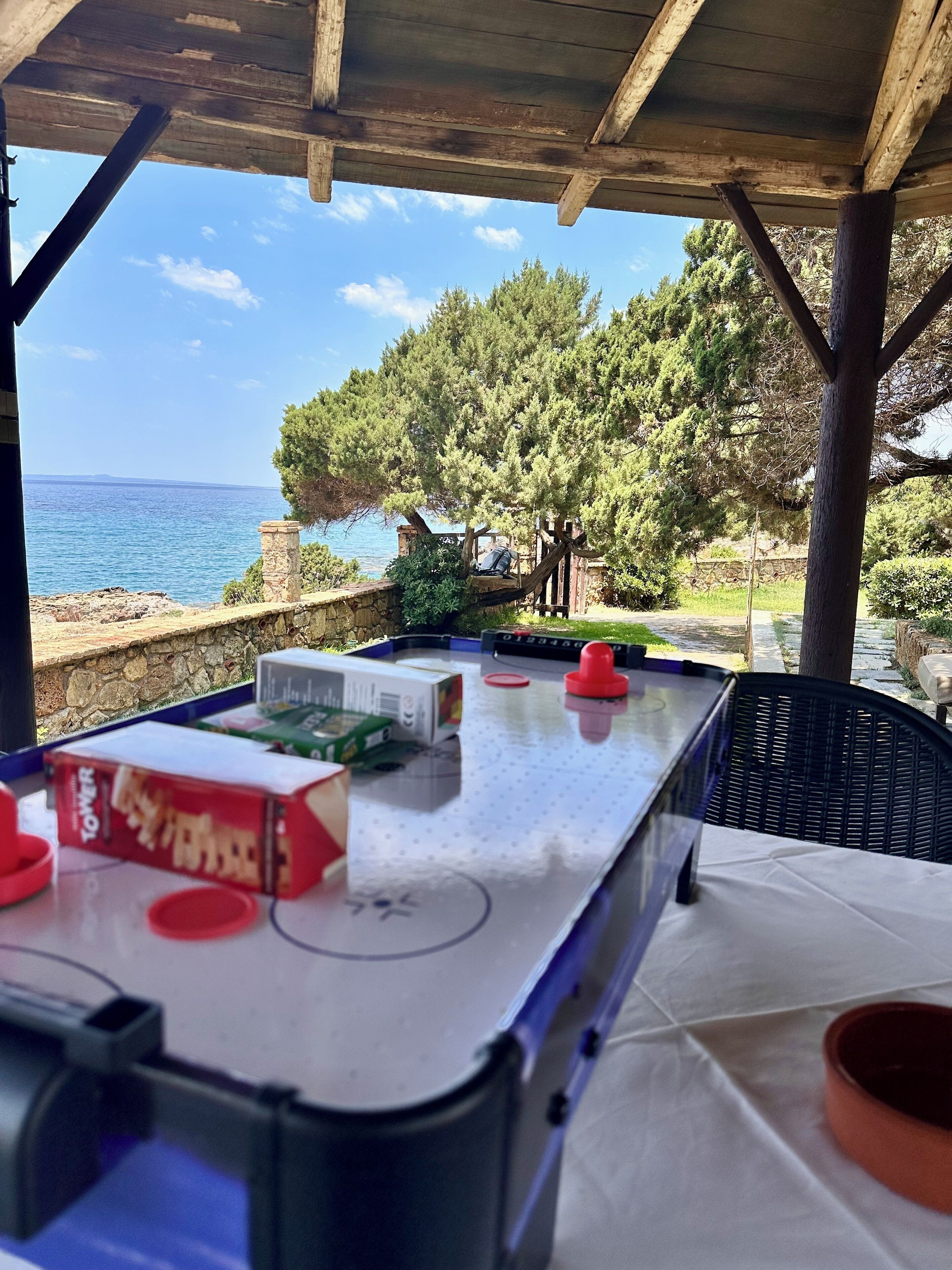 Uma mesa de bilhar com tacos e uma caixa de bolas de bilhar, uma parede de pedra e vista para o mar e árvores.