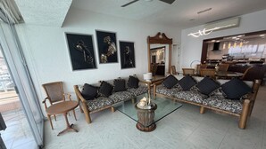 Una sala de estar con un sofá, sillas, una mesa de centro de vidrio y una cocina al fondo.