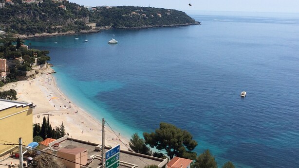 Uitzicht op de kust met een strand, boten en een bord met de richting naar Nice en Menton.