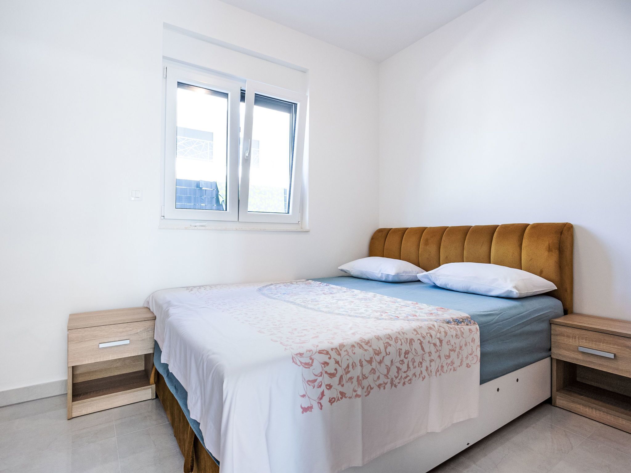 A bedroom with a bed, two bedside tables, a window, and a view of buildings outside.