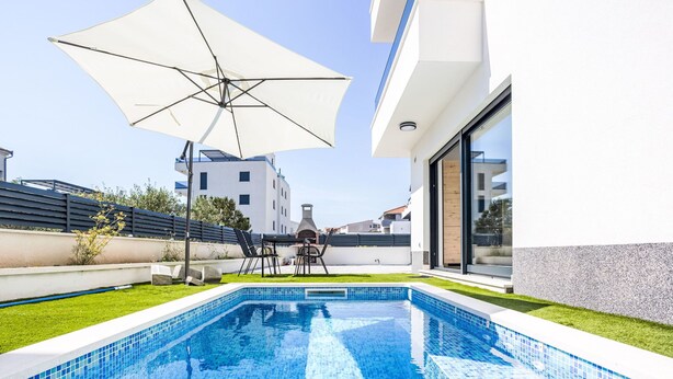 A swimming pool with a tiled edge, a patio area with a table and chairs, and a large umbrella. - Sunny Pool by Interhome (Trogir/Okrug Gornji)