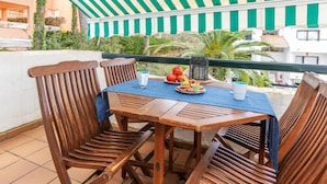 Ein Holztisch mit Stühlen unter einer Streifenmarkise, ein Teller mit Essen und ein Getränk auf einem Balkon mit Blick auf Gebäude und Grünflächen.