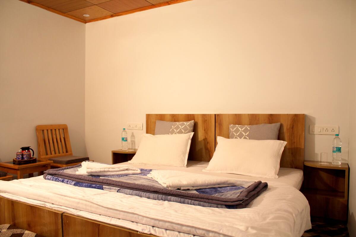 Una cama bien hecha con cabecera de madera, dos mesitas de noche con lámparas, una silla de madera y una pequeña mesa con una tetera y tazas.