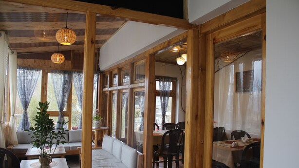 A cozy room with a wooden ceiling, a sofa, a dining table with chairs, and large windows with curtains. - Karma Lodge (Leh)
