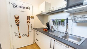 A kitchen with a wooden floor, white cabinets, a black countertop, a stainless steel sink, a microwave above the counter, and a door with a sign that reads "Bonjour".