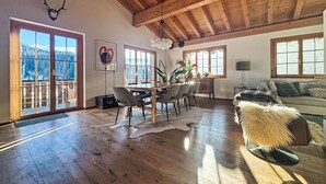 Een ruime woonkamer met een houten plafond, een eettafel met stoelen, een bank en een groot raam met uitzicht op een besneeerd landschap.