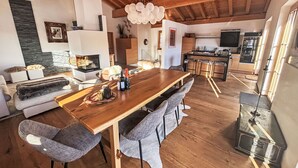 Een ruime woonkamer met een houten eettafel, grijze fauteuils, een open haard en een keuken op de achtergrond.