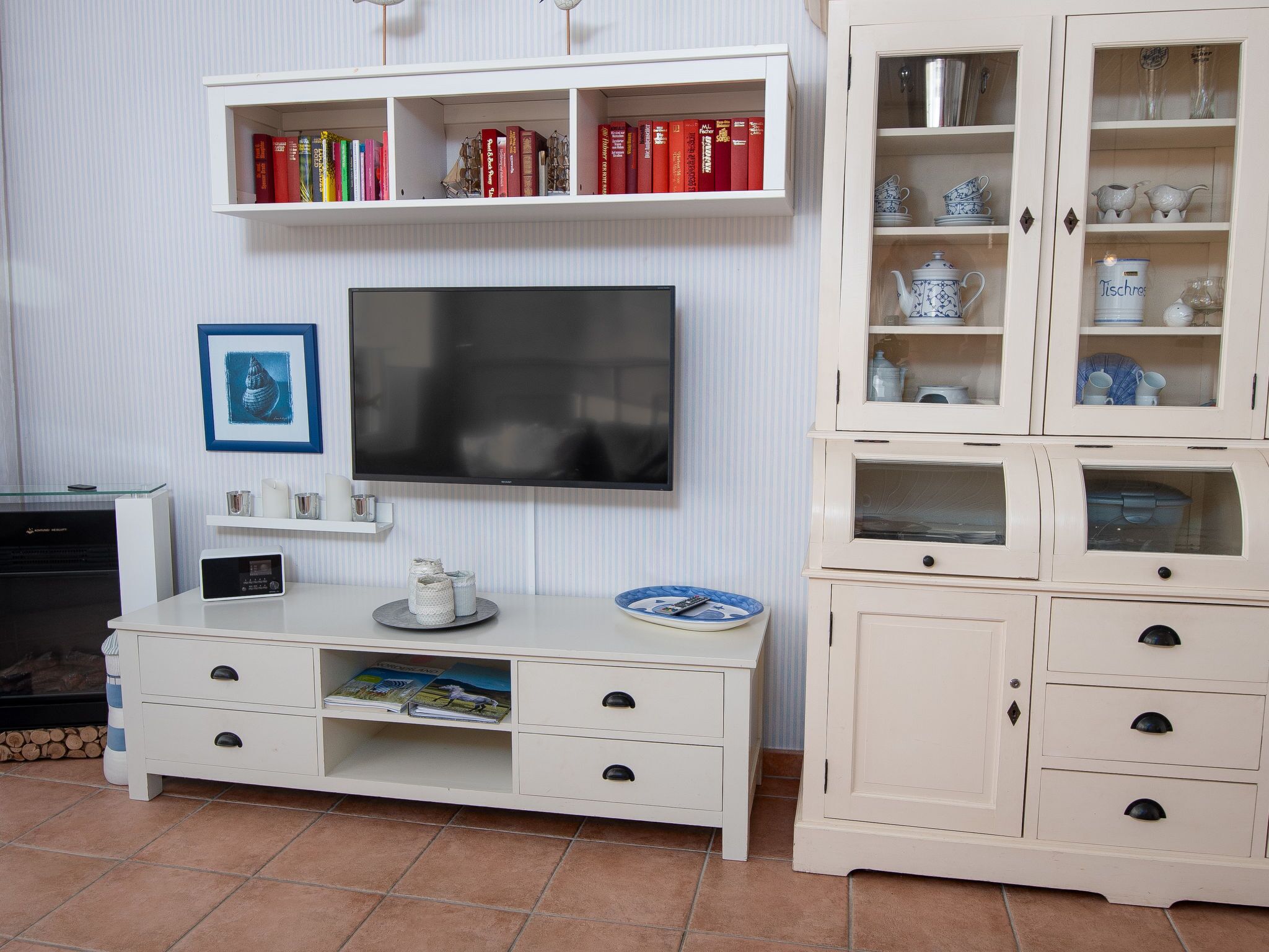 Un salón con televisor de pantalla plana, un mueble blanco para entretenimiento con libros, un cuadro enmarcado y un armario con puertas de vidrio que contiene utensilios de cocina.