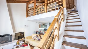 Un interior moderno con una escalera de madera que sube a una zona tipo loft, un sofá con cojín escocés, y una cocina al fondo.