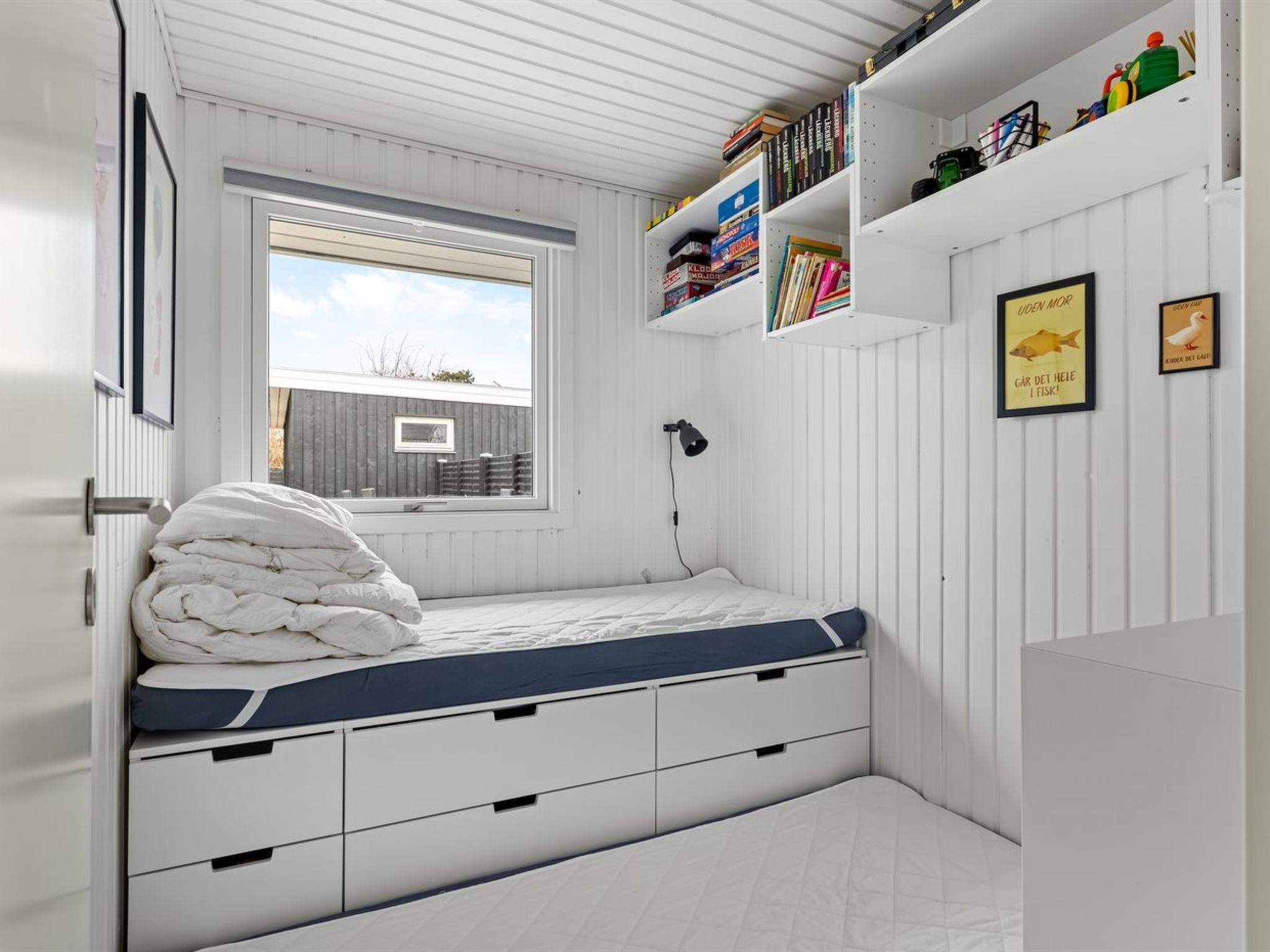 A compact bedroom with a bed, built-in storage, a window, and a bookshelf.