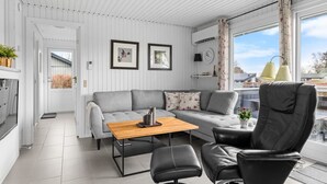 A modern living room with a grey sofa, a wooden coffee table, a black leather chair, and a dining table. There are two framed pictures on the wall, a clock, and a window with curtains. - "Bjart" - 150m from the sea by Interhome (Hesselager)
