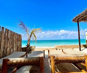 A beach with wooden lounge chairs and a thatched roof structure. - Aquelia Rose Hotel (Jambiani)