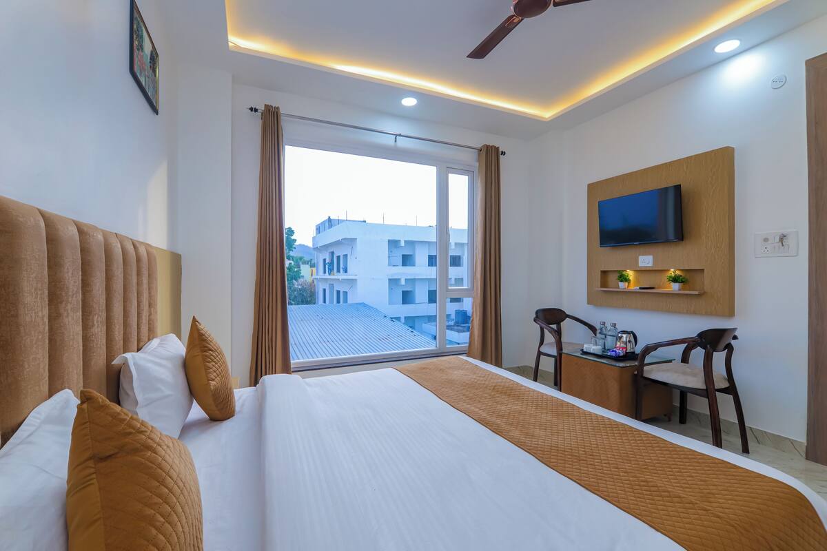 A hotel room with a large bed, a desk with a chair, a television, and a view of buildings through the window.