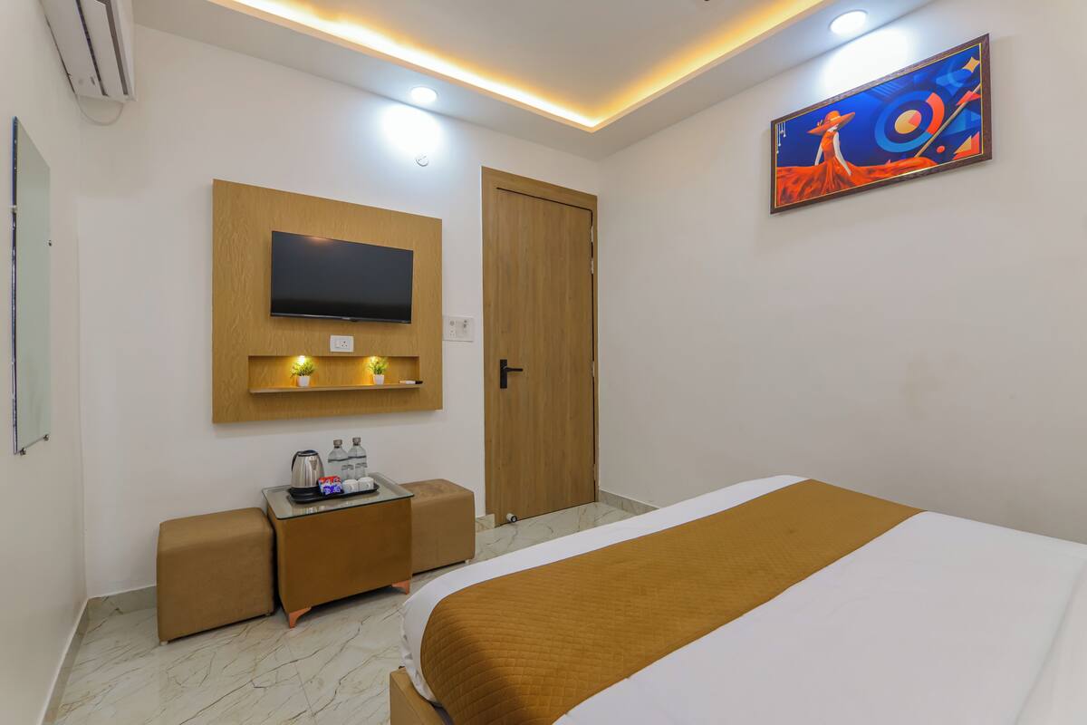 A hotel room with a bed, a TV mounted on a wooden panel, a small desk with a coffee maker, and a painting on the wall.
